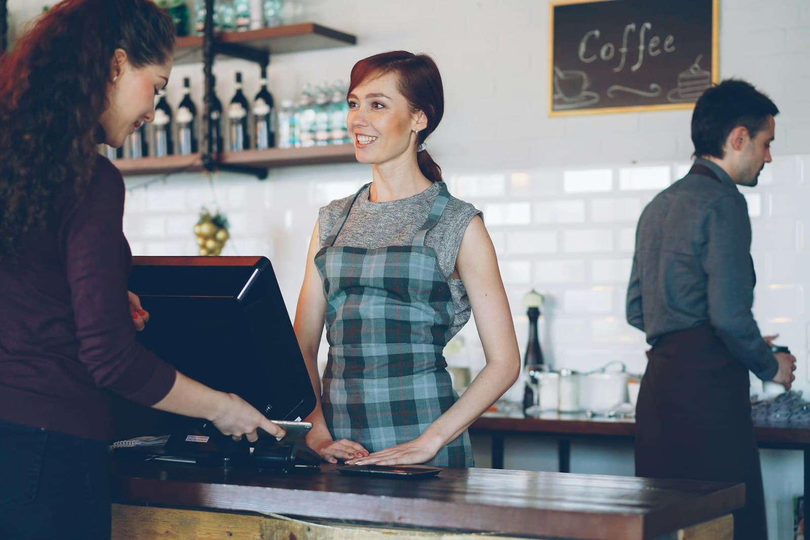 Une cliente règle sa commande sur le terminal de caisse d'un café, face à une serveuse souriante, pendant qu'un barista travaille au comptoir