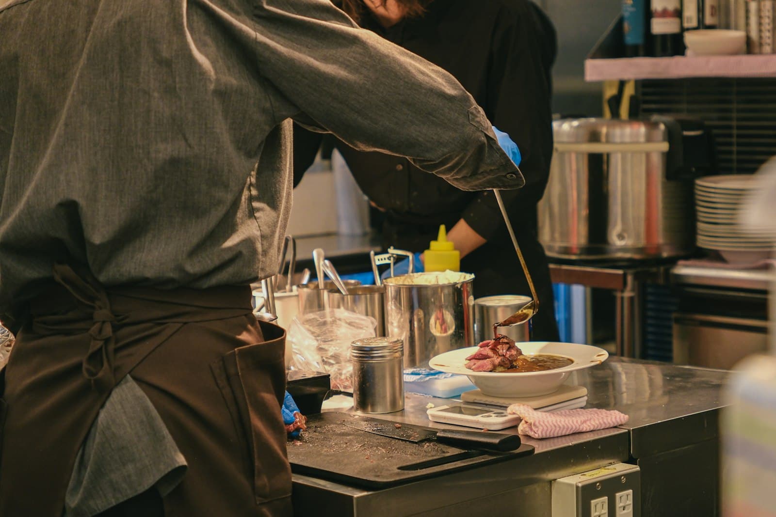 Deux cuisiniers dressent un plat de viande nappé de sauce sur un plan de travail en inox, entre marmites et piles d'assiettes