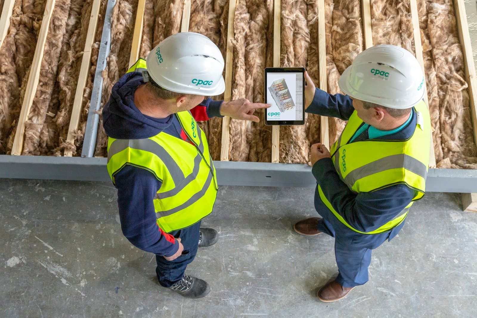 Deux compagnons en gilet fluorescent et casque consultent une maquette numérique sur une tablette, devant une ossature bois isolée