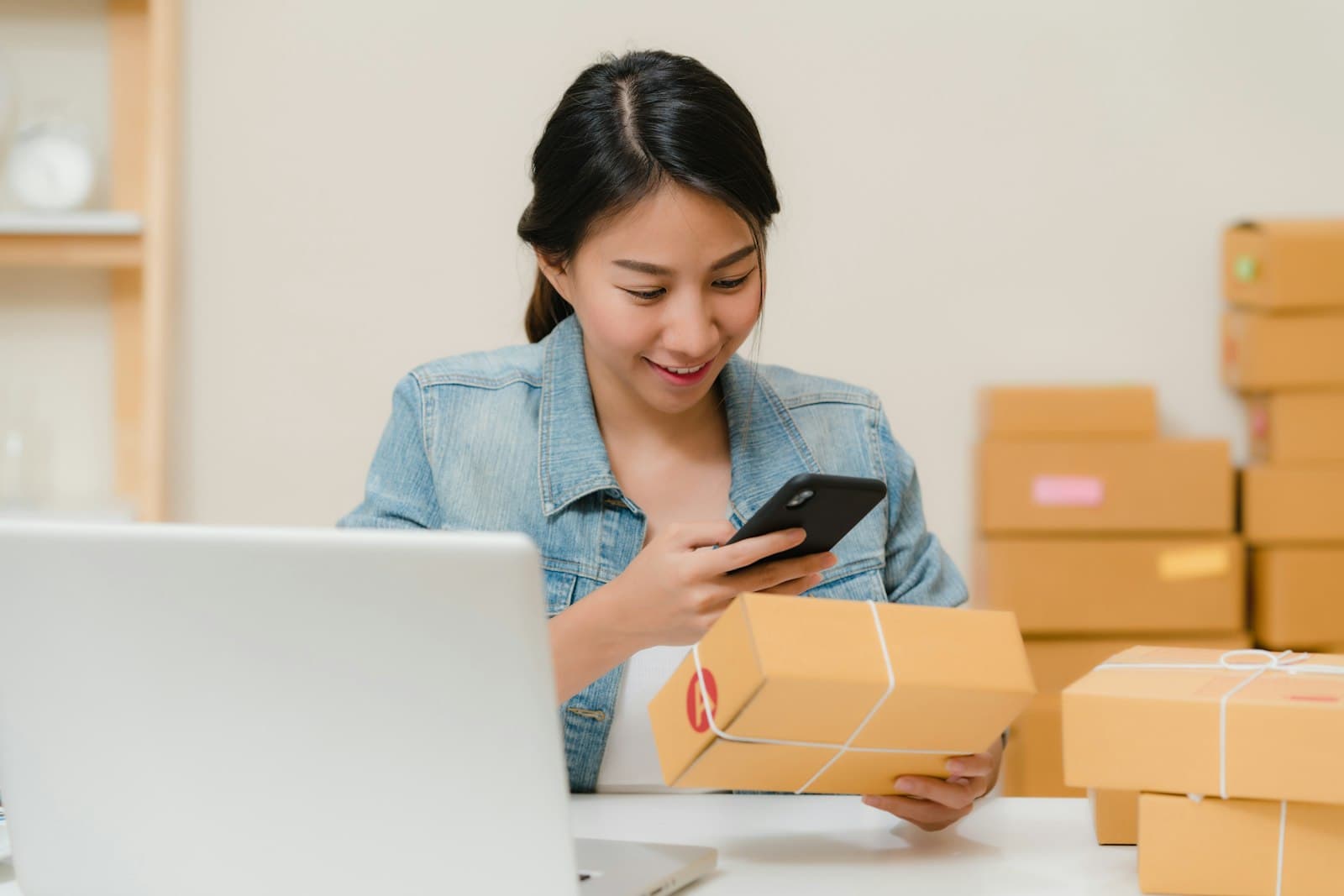 Une commerçante en ligne scanne un colis avec son téléphone devant son ordinateur portable, entourée de cartons d'expédition