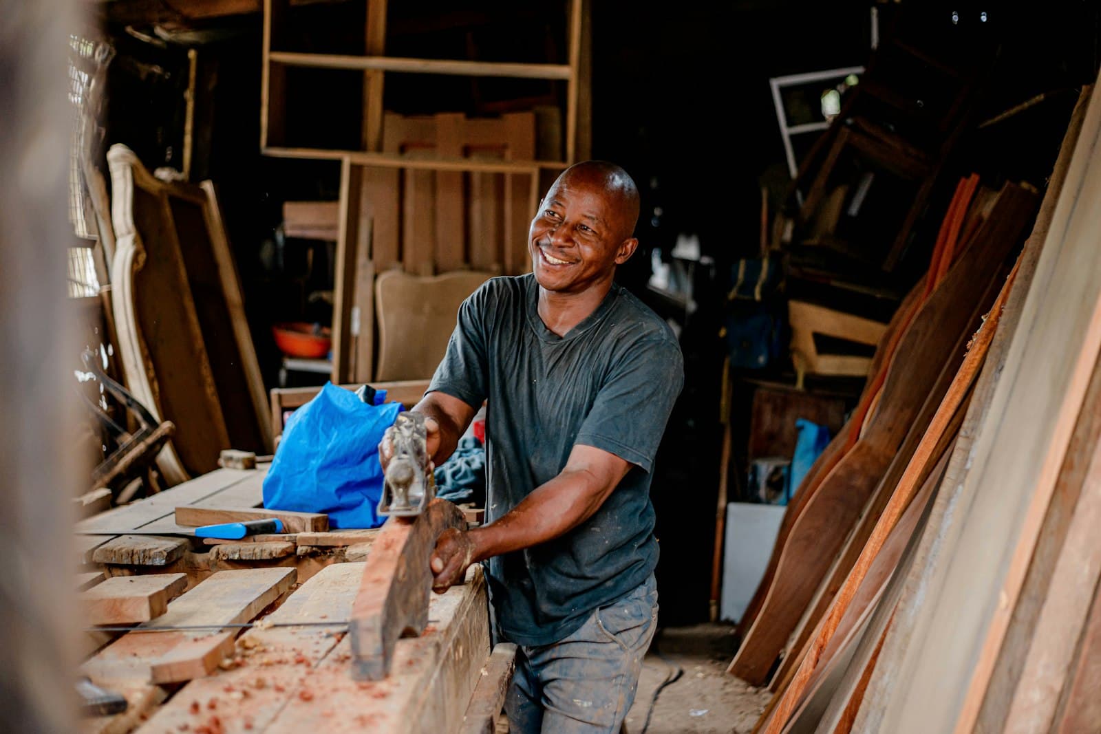 Un artisan menuisier souriant rabote une planche dans son atelier, entouré de bois de construction
