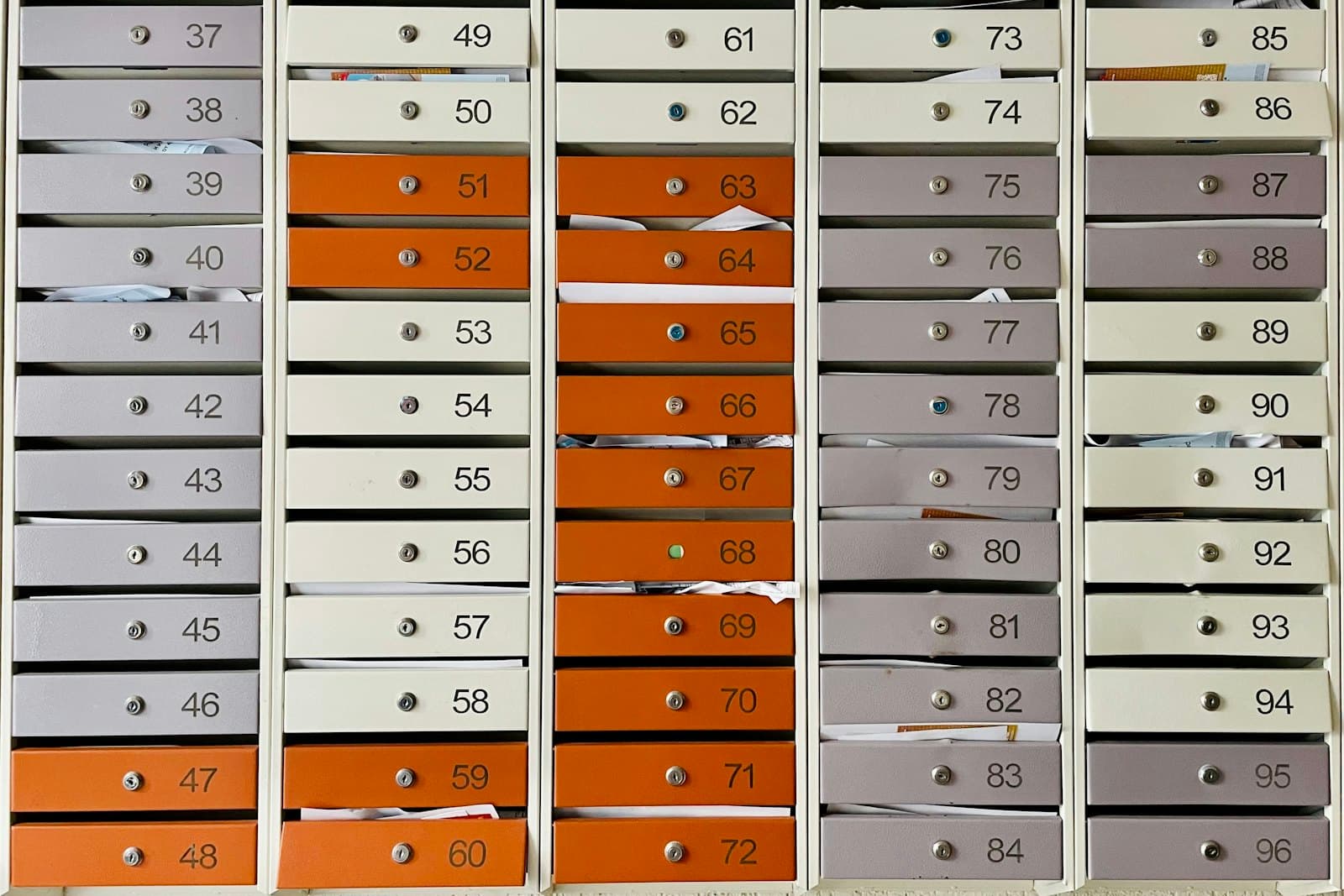 Un mur de boîtes aux lettres métalliques numérotées, en orange, crème et gris, dont certaines débordent de courrier