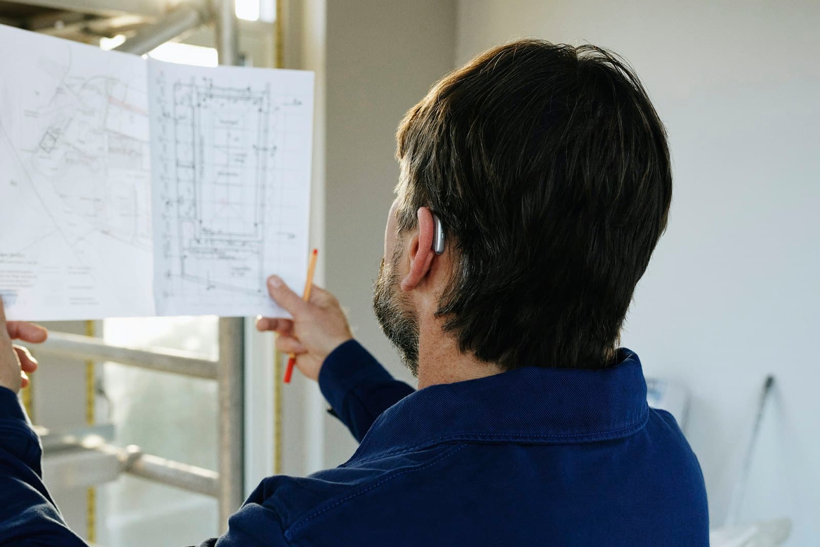 Une personne en veste de travail bleue, vue de dos, examine un plan de bâtiment déplié, un crayon de charpentier sur l'oreille