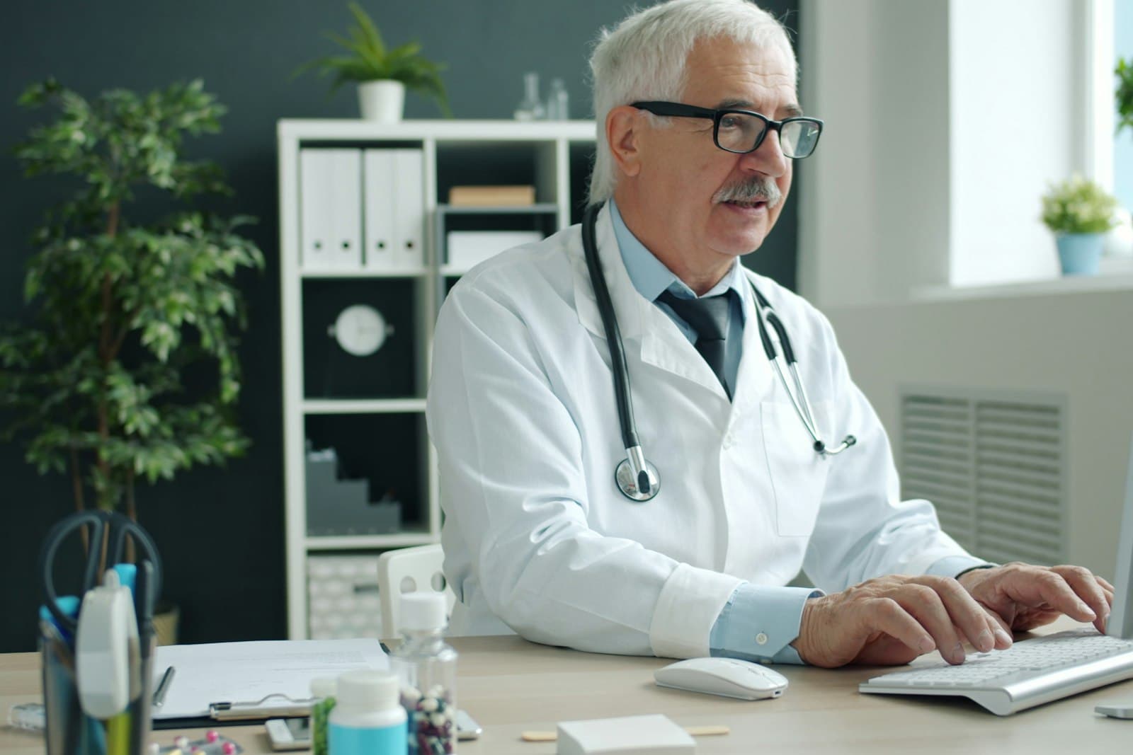 Un médecin en blouse blanche, stéthoscope autour du cou, saisit une consultation sur le clavier de son ordinateur dans son cabinet