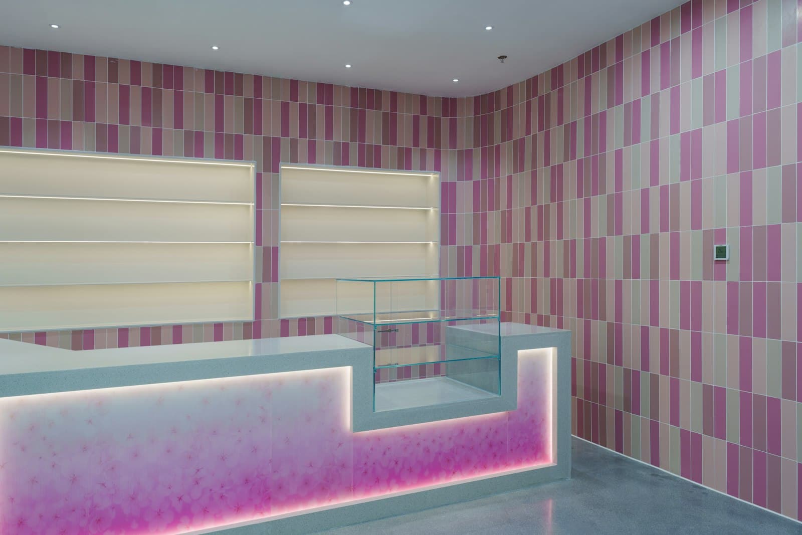 Comptoir d'une boutique neuve aux murs carrelés de rose, avec une vitrine vide et des étagères éclairées