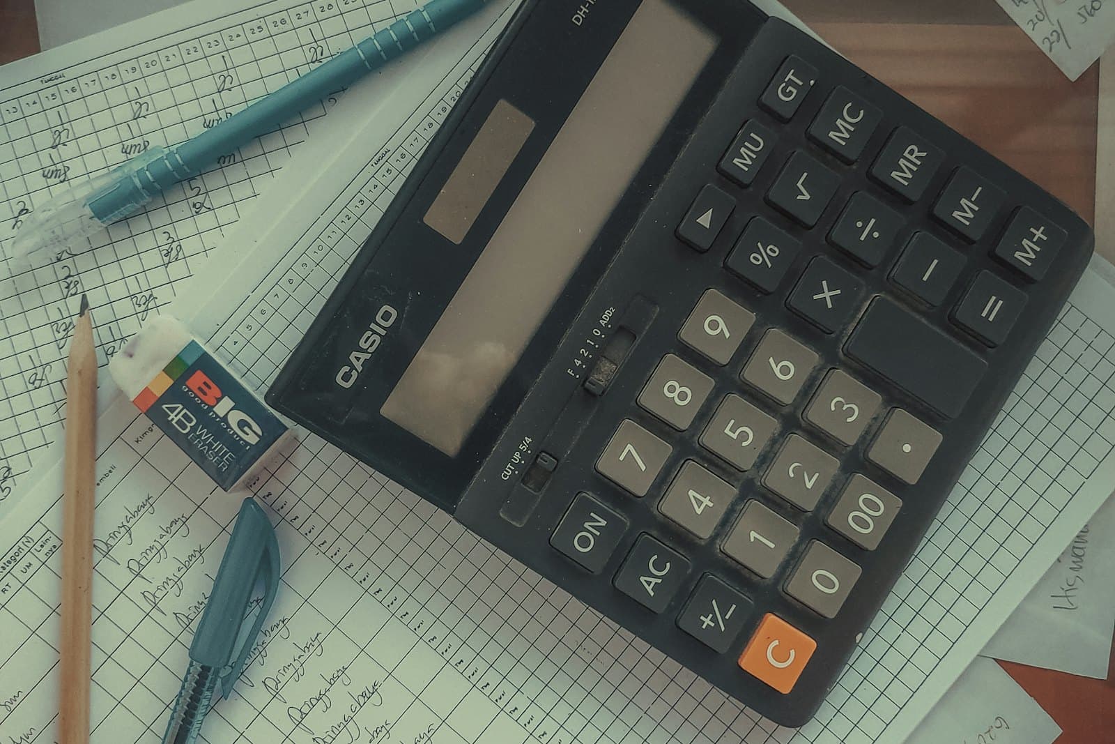 Une calculatrice posée sur des feuilles de suivi quadrillées remplies à la main, entourée de stylos, d’un crayon et d’une gomme