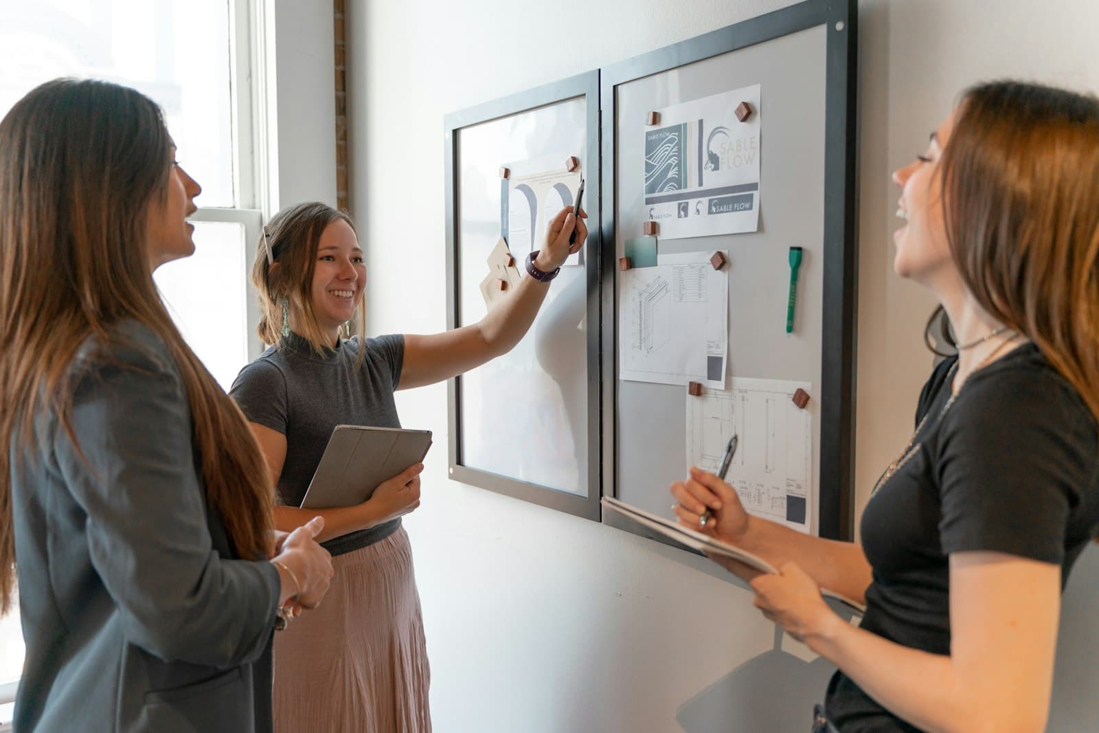 Trois collègues discutent devant un tableau de projet mural couvert de plans et de documents épinglés