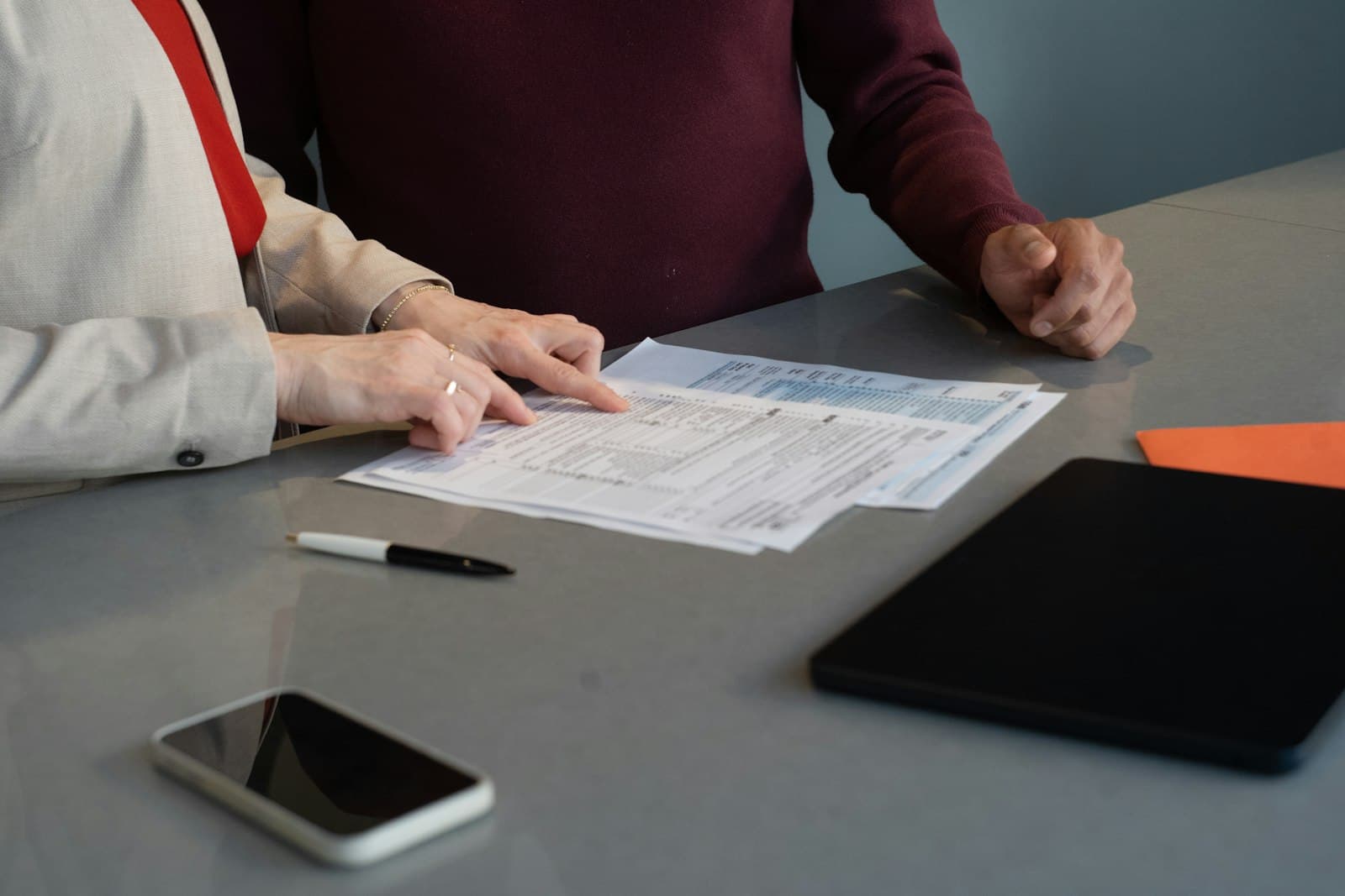 Deux personnes examinent des documents administratifs imprimés sur une table de réunion, un stylo et une tablette posés à côté