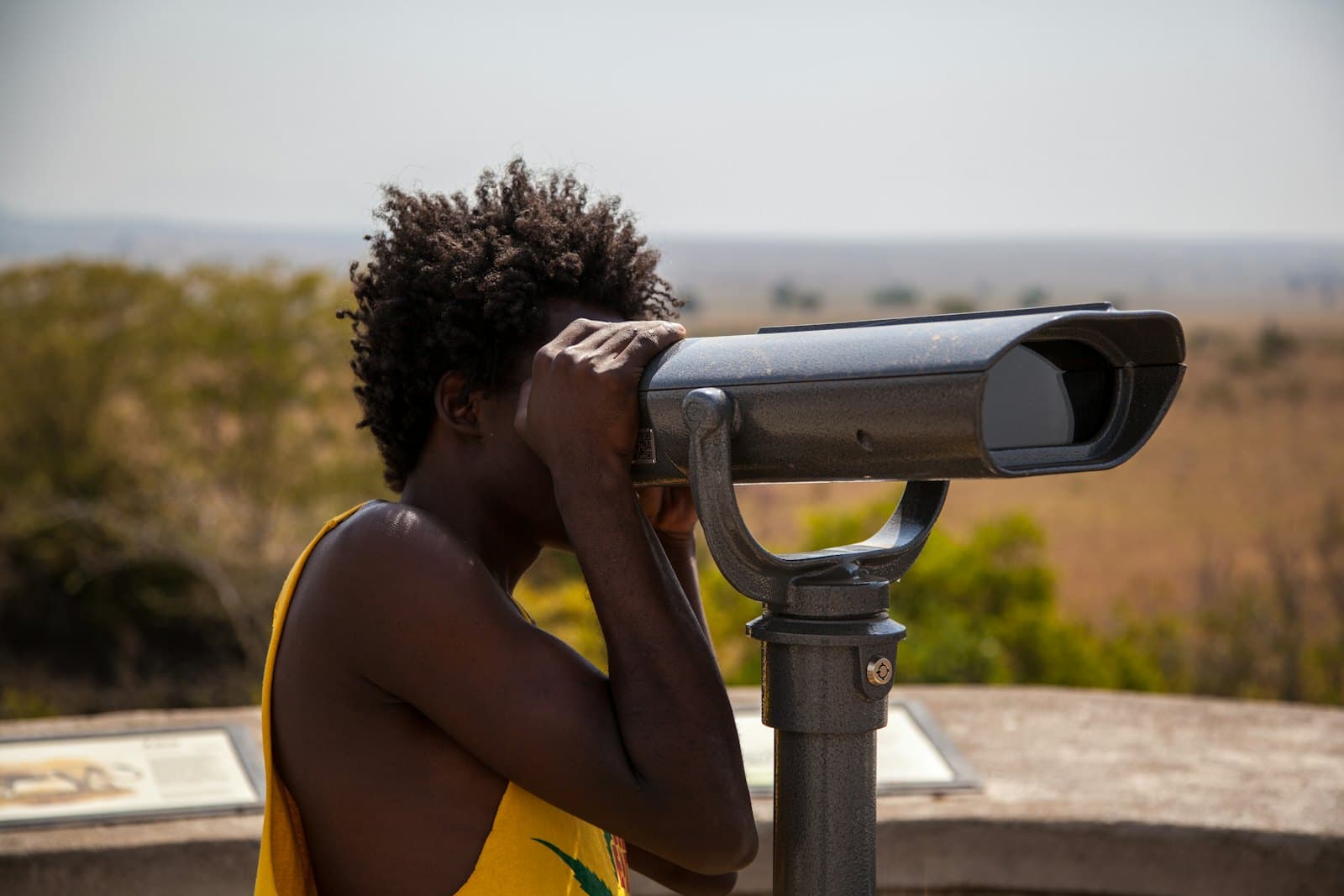 Une personne en débardeur jaune observe un paysage de savane à travers une longue-vue payante installée sur un belvédère